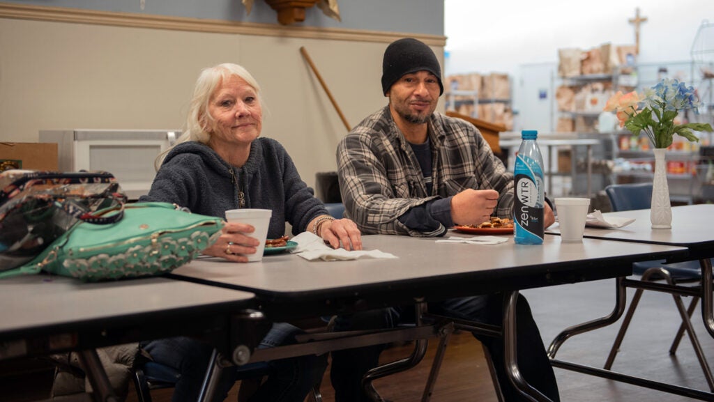 A woman and a man dine at a Catholic Charities congregate dining center. She has white hair and wears a hoodie, he has a plaid shirt on and a dark knit cap.