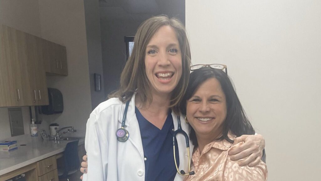 New mom Sarah stands in her nurse's scrubs with her arm around Chasity, adoption program director. They're smiling and look proud and happy.